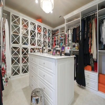 Luxury walk‑in closet with white custom cabinetry, glass‑front wardrobe doors, center island drawers, and chandelier lighting in a Long Island home